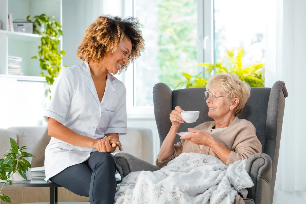 A caregiver sitting with an older woman as she drinks tea in her living room